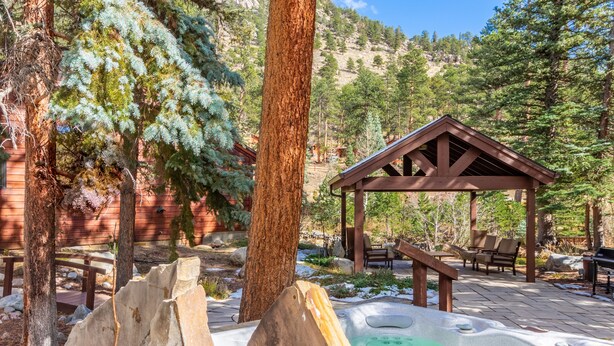 A hot tub surrounded by wooden benches and a gazebo in a forested area with a mountain backdrop. - Boulder Brook on Fall River (Estes Park)