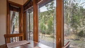 A wooden dining table with two woven placemats and a chair, set against a backdrop of a forested landscape visible through large glass doors. - Boulder Brook on Fall River (Estes Park)