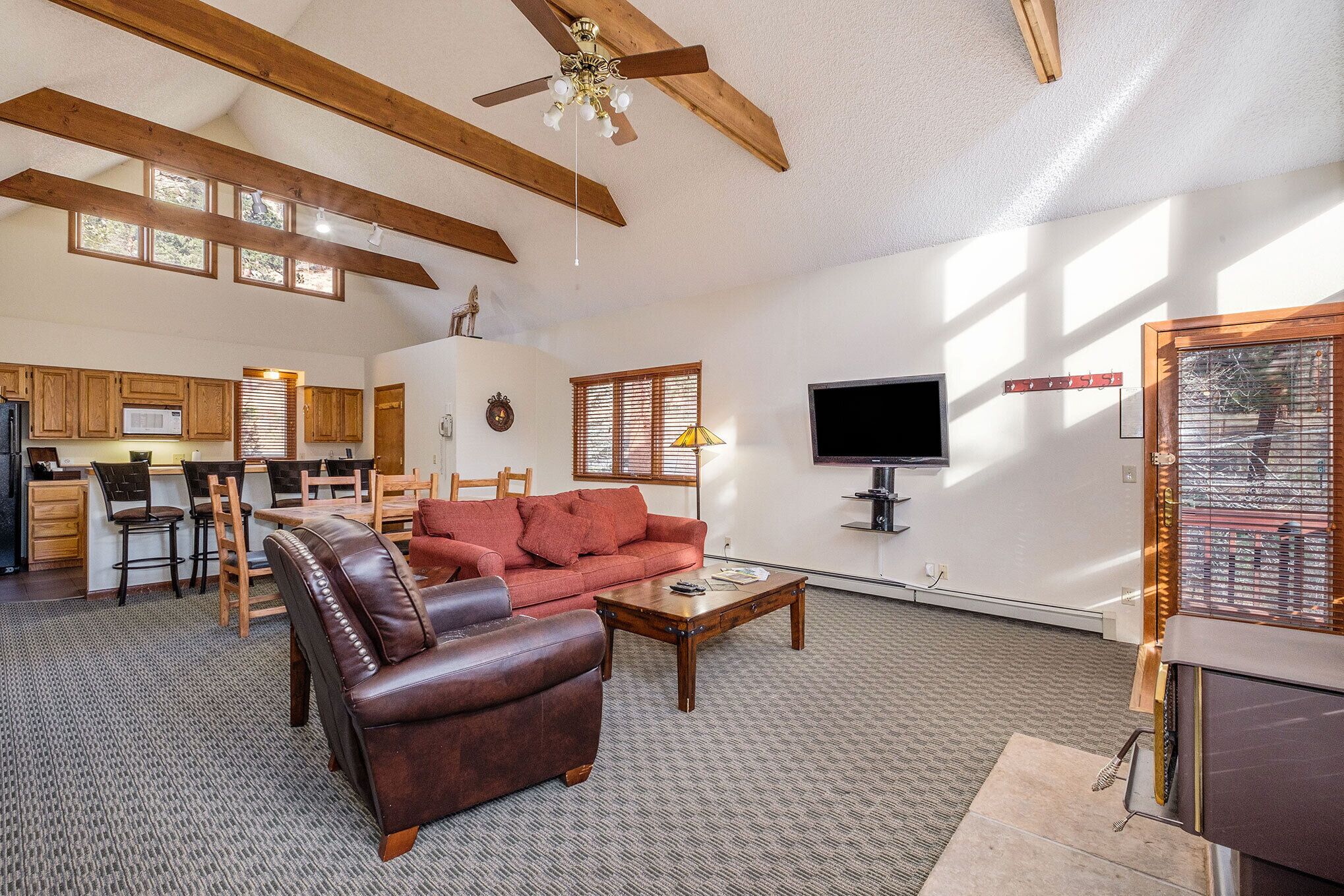 A spacious living room with a leather sofa, a coffee table, and a TV. The room has wooden beams on the ceiling and a kitchen in the background.