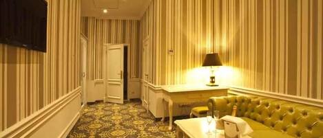 Habitación de hotel con papel tapiz a rayas, un sofá verde, una mesa con lámpara y una alfombra estampada.