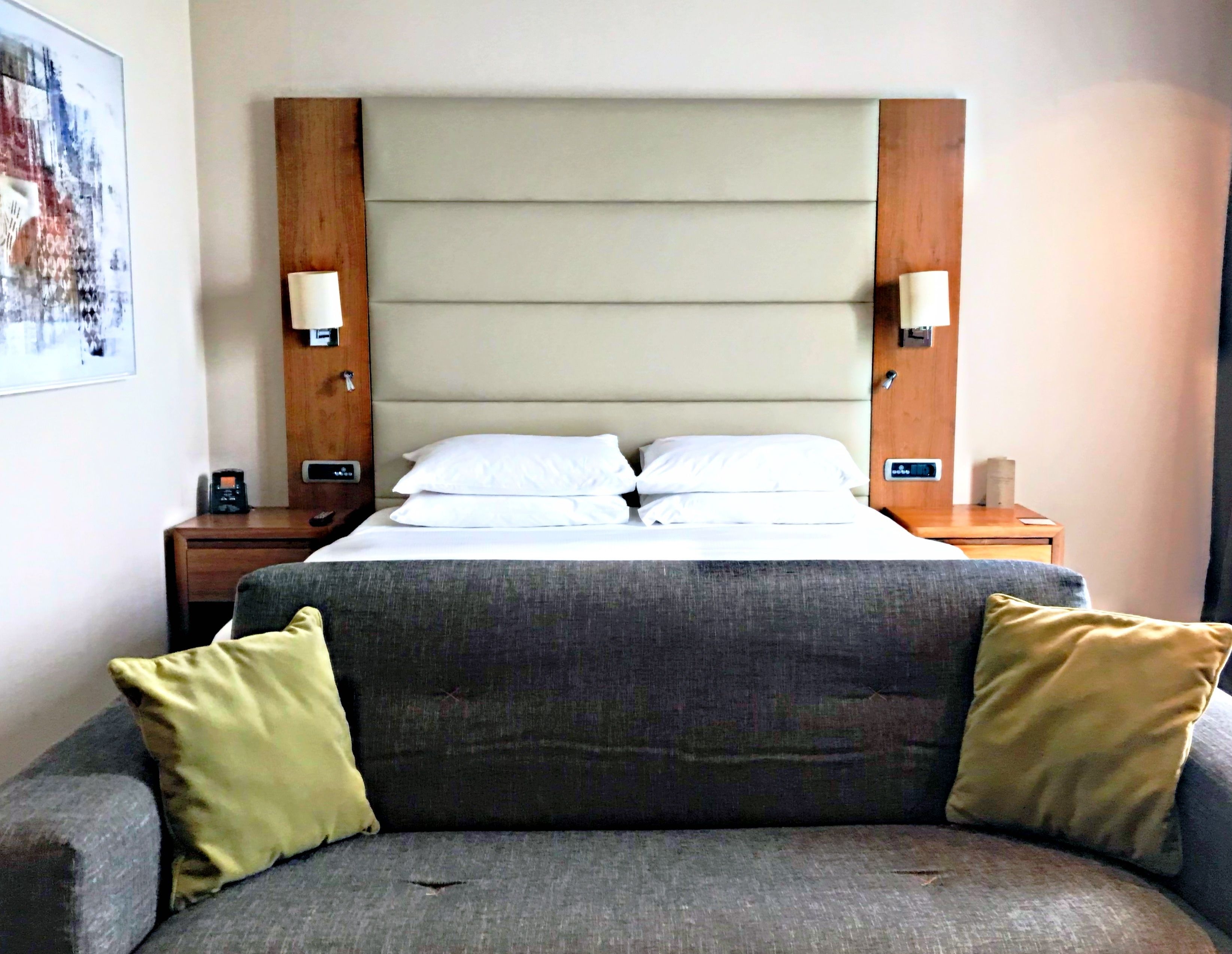 A hotel room with a bed, two bedside tables, a nightstand, a gray sofa, and a painting on the wall.