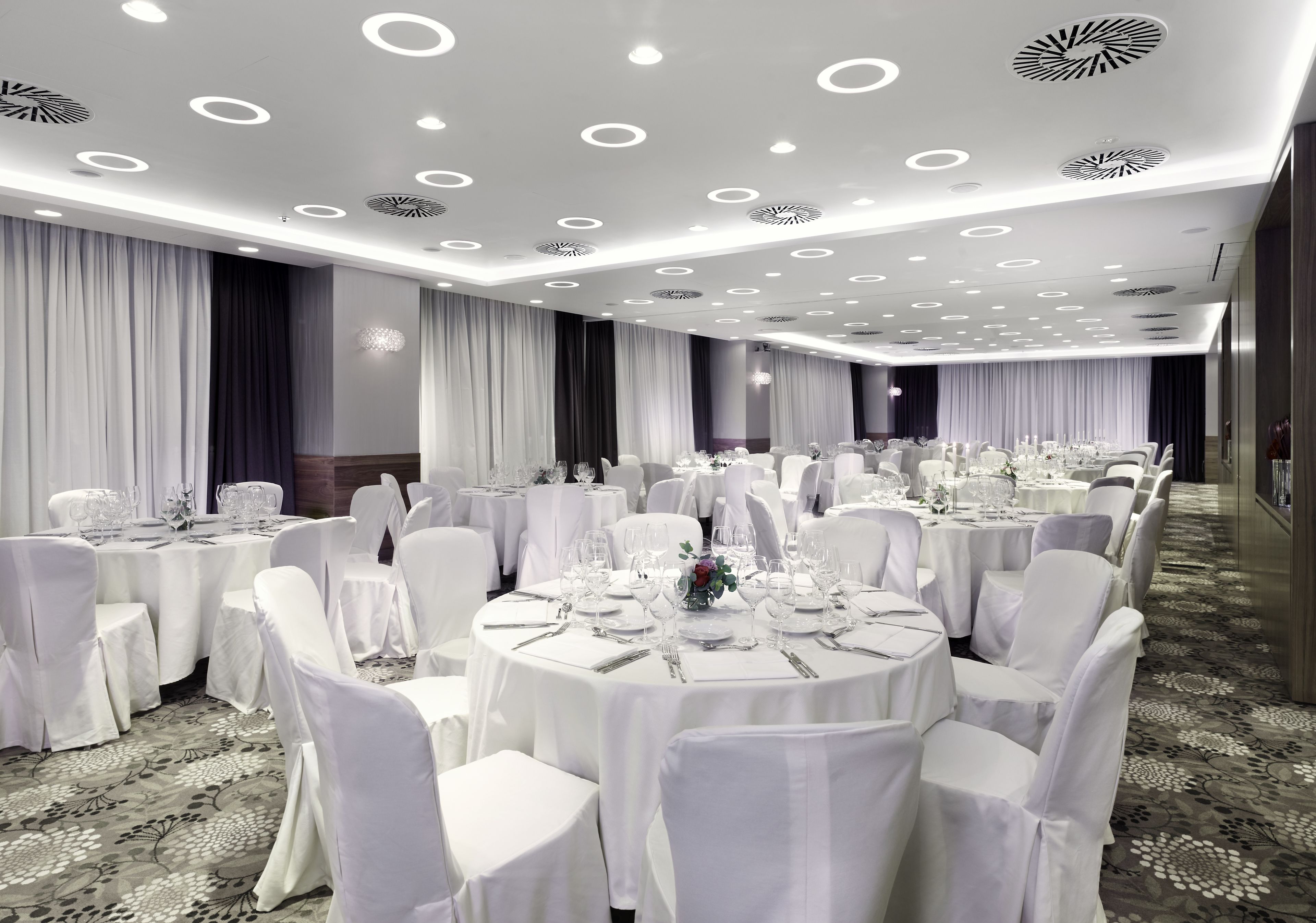 A banquet hall with round tables, white tablecloths, and chairs, featuring a modern ceiling design with circular lights.