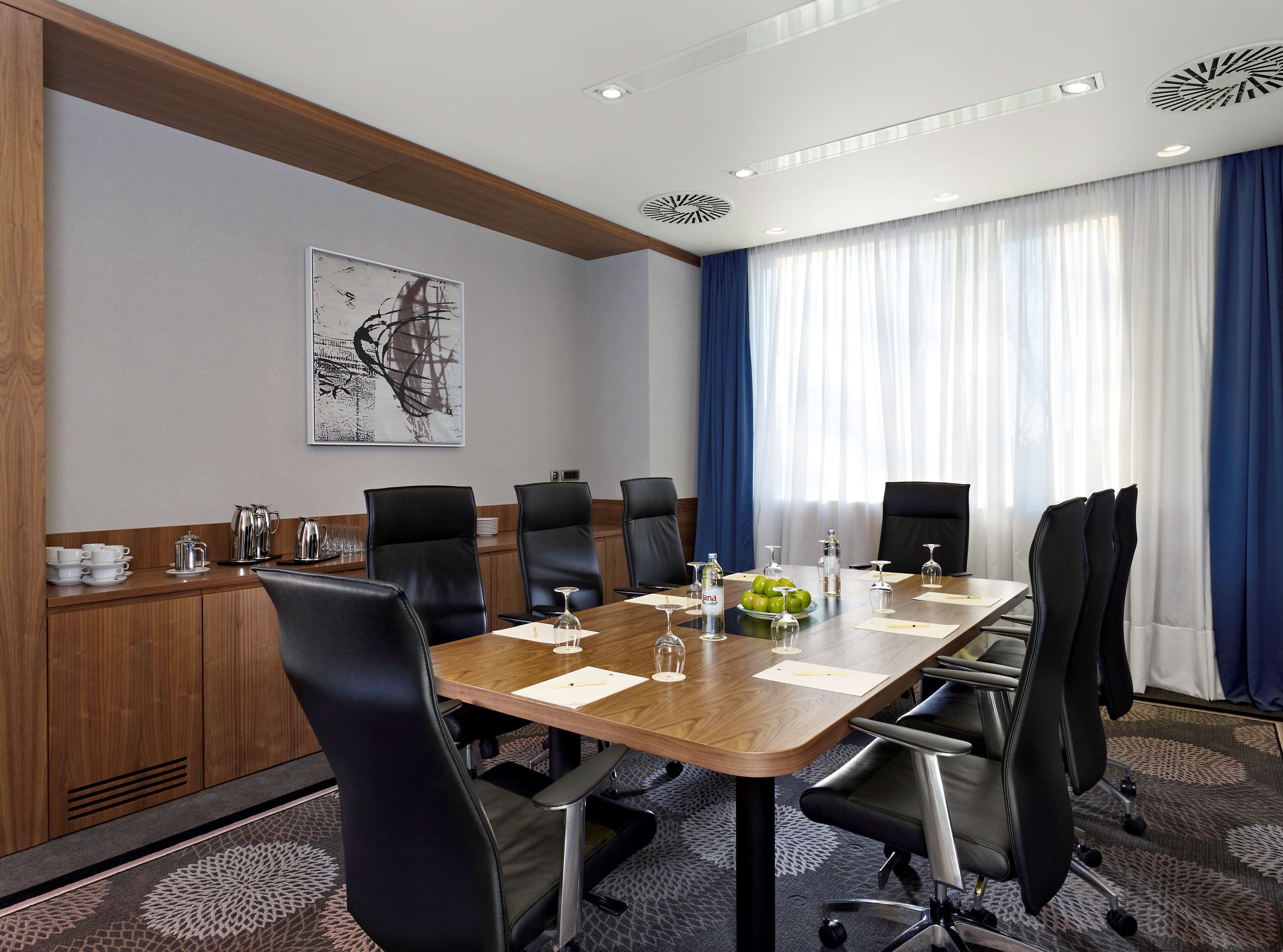 A conference room with a long wooden table, black chairs, and a large window with blue curtains.
