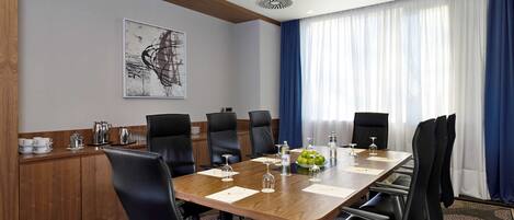 A conference room with a long wooden table, black chairs, and a large window with blue curtains.