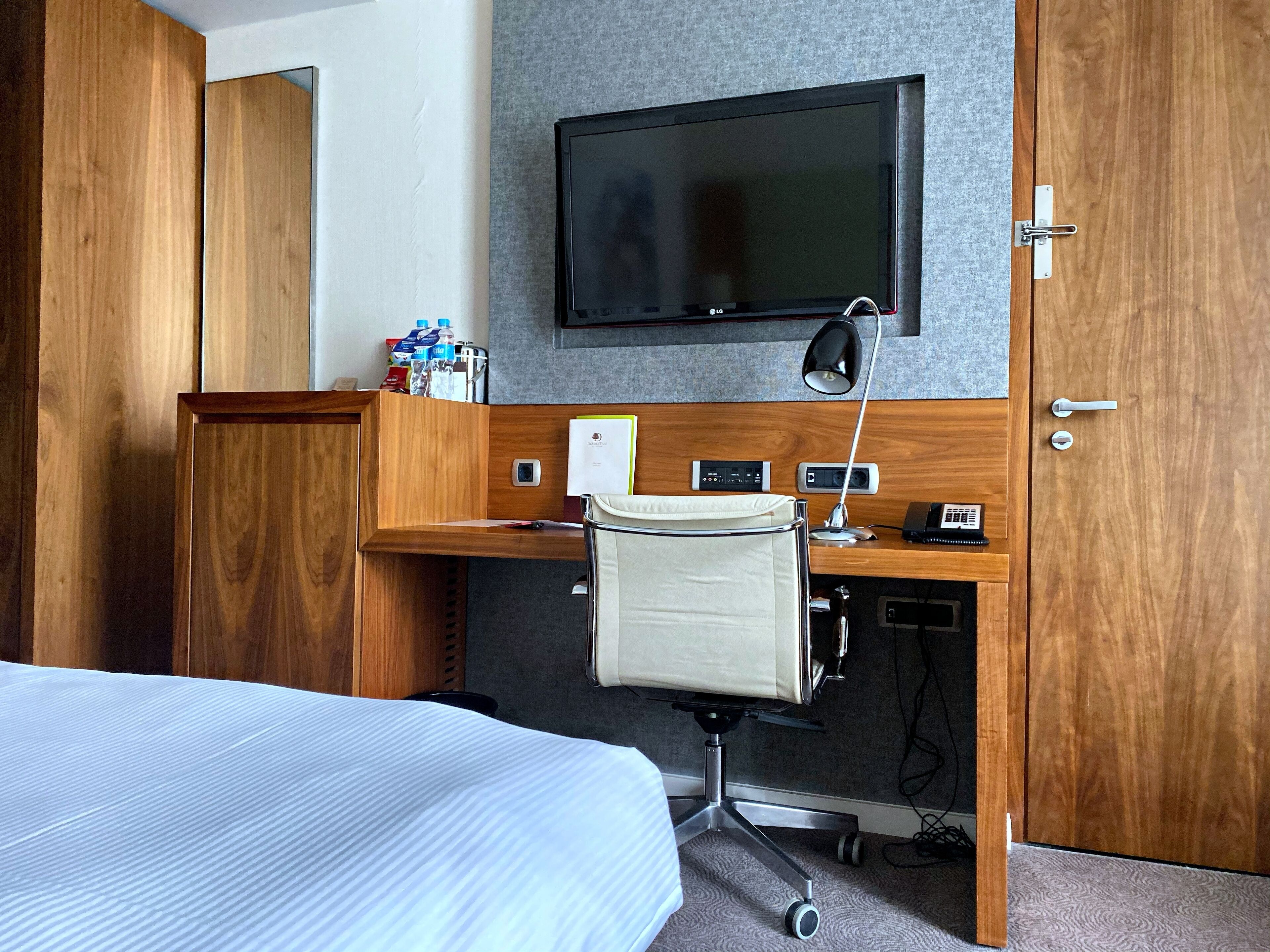 A hotel room with a bed, a desk with a chair, a TV mounted on the wall, and a wooden wardrobe.