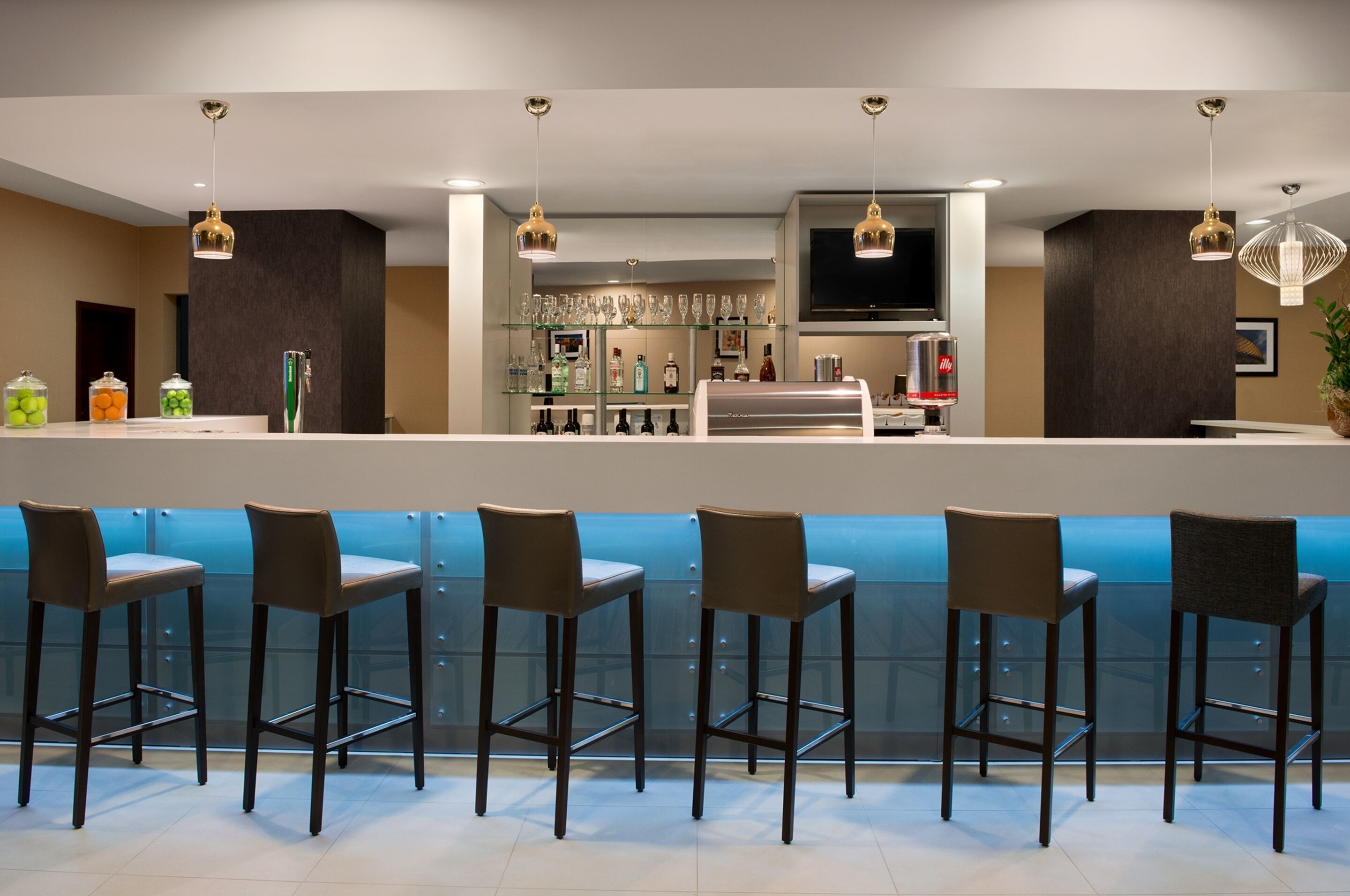Eine moderne Bar mit hohen Hockstühlen, einer Theke mit Fernseher und verschiedenen Flaschen und Gläsern.