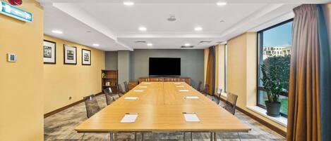 Uma sala de conferências com uma mesa comprida, cadeiras, uma tela de TV e uma janela com cortinas.