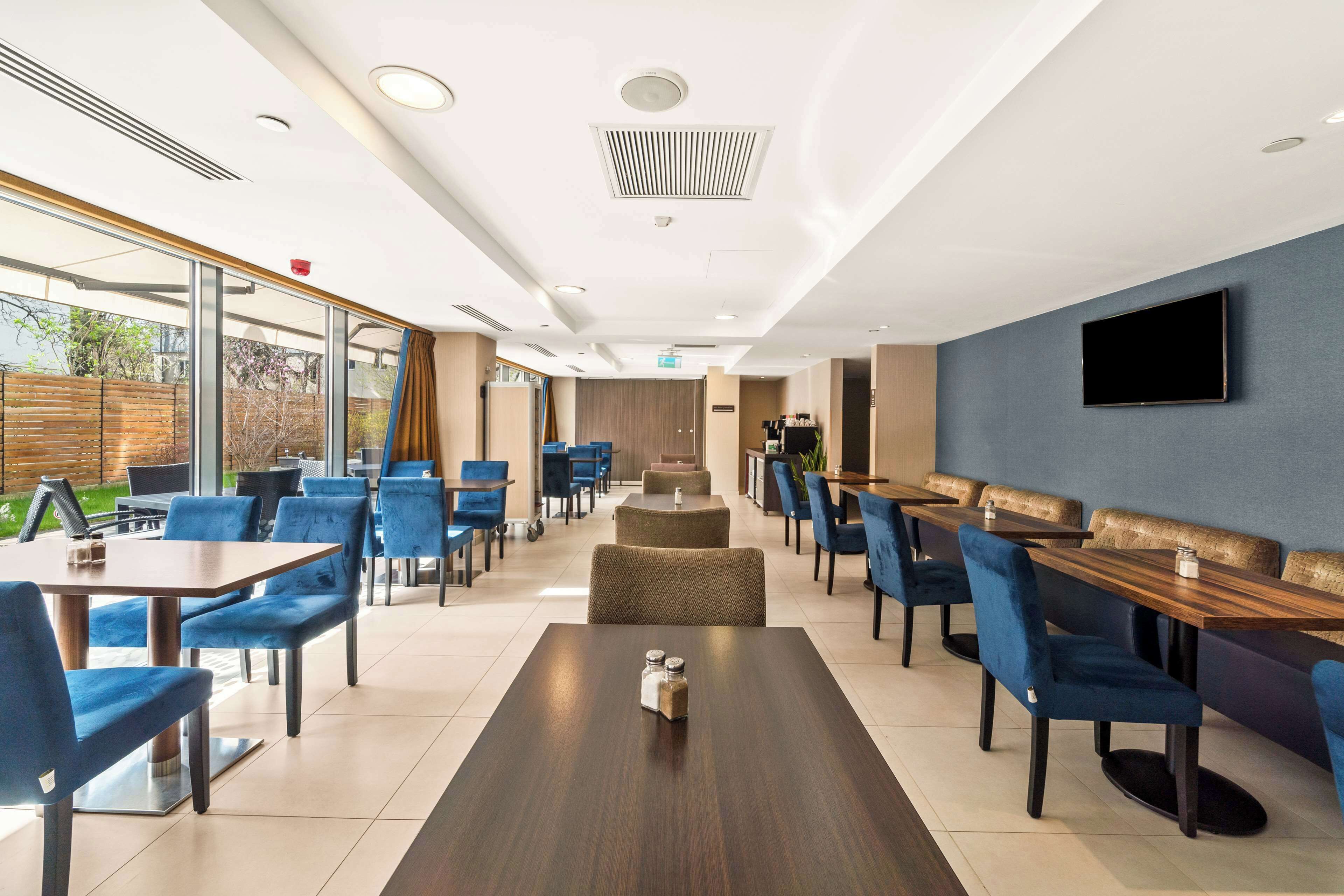 A modern dining area with tables and chairs, a flat-screen TV mounted on the wall, and a view of the outdoors through large windows.