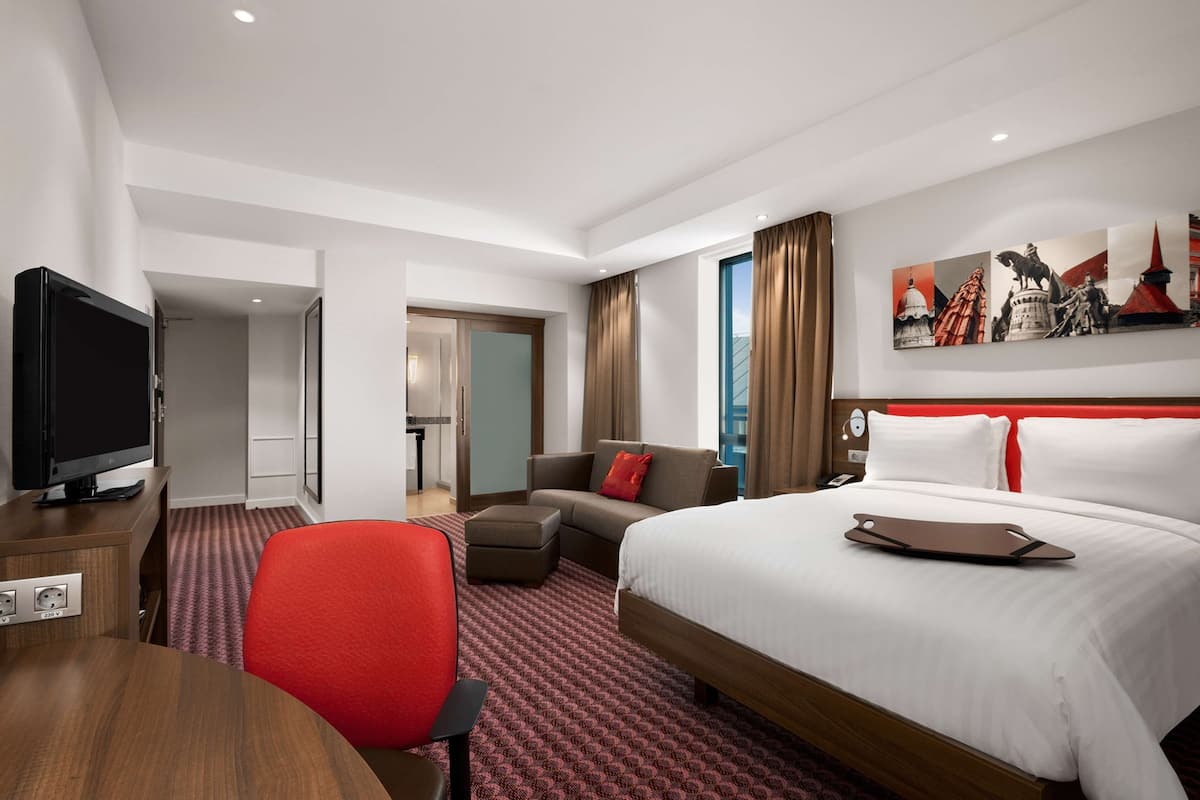 Une chambre d’hôtel avec un lit, une télévision, un bureau, une chaise rouge, un canapé et un tableau au mur.