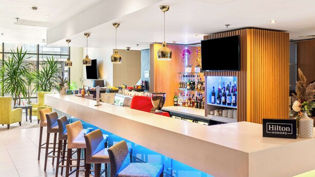 A modern bar area with a long counter, multiple bar stools, a television mounted on the wall, and a variety of bottles and glasses on shelves. - Hampton by Hilton Cluj-Napoca (Cluj-Napoca)