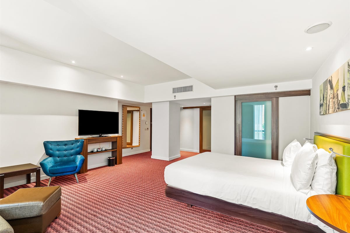 Une chambre d’hôtel avec un lit, un fauteuil bleu, une table de chevet en bois, une télévision et un sol recouvert d’une moquette rouge.