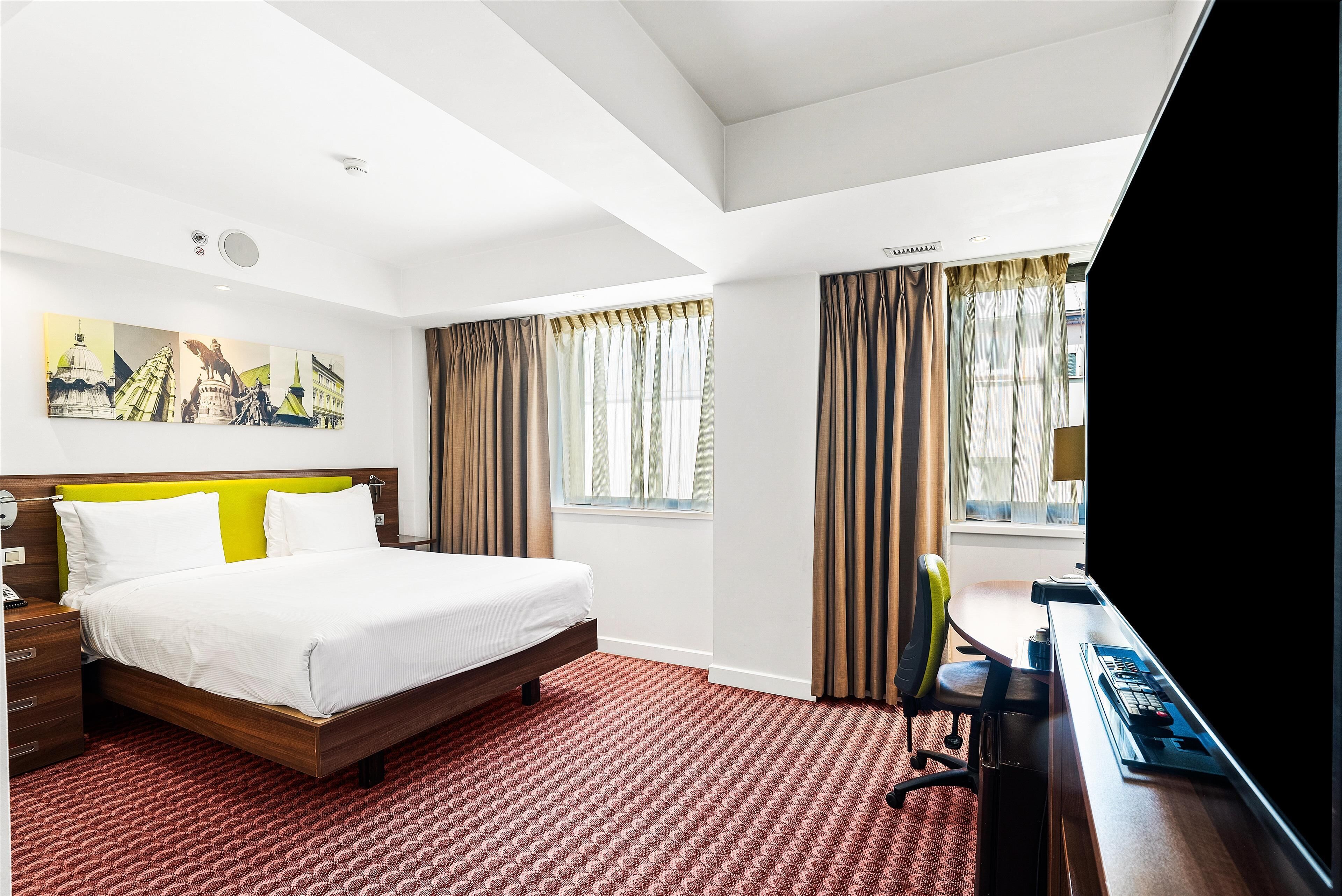 A hotel room with a bed, a desk with a computer, a chair, and a large window with curtains.