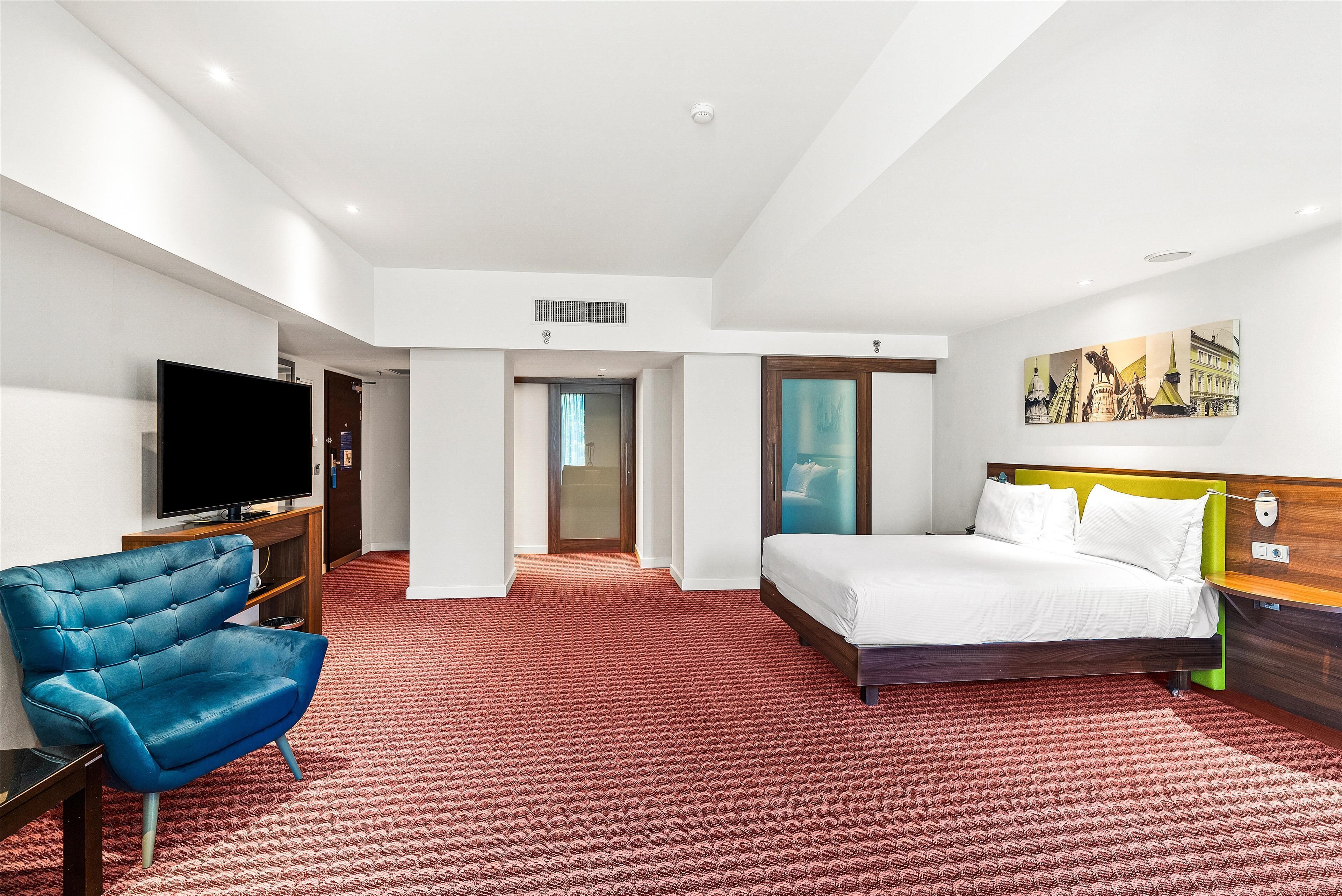 Quarto de hotel com cama, televisão, poltrona azul e tapete vermelho estampado.