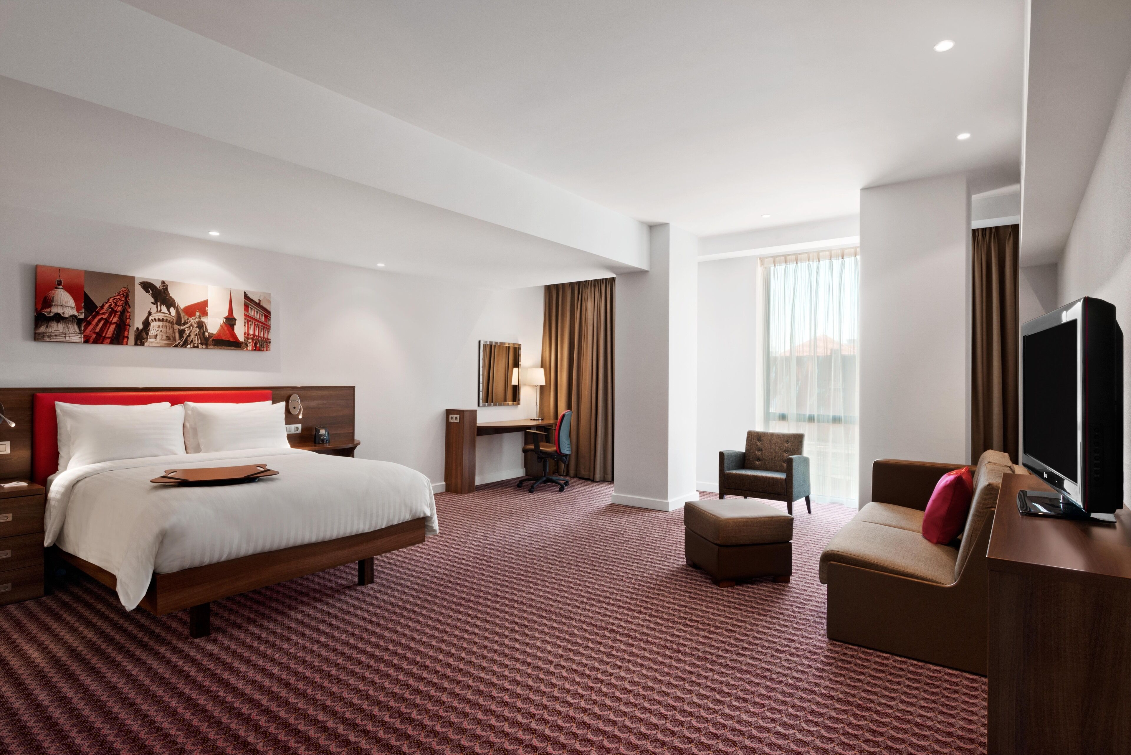 Ein Hotelzimmer mit einem großen Bett, einem Schreibtisch, einem Sessel, einem Fernseher und einem Fenster mit Vorhängen.