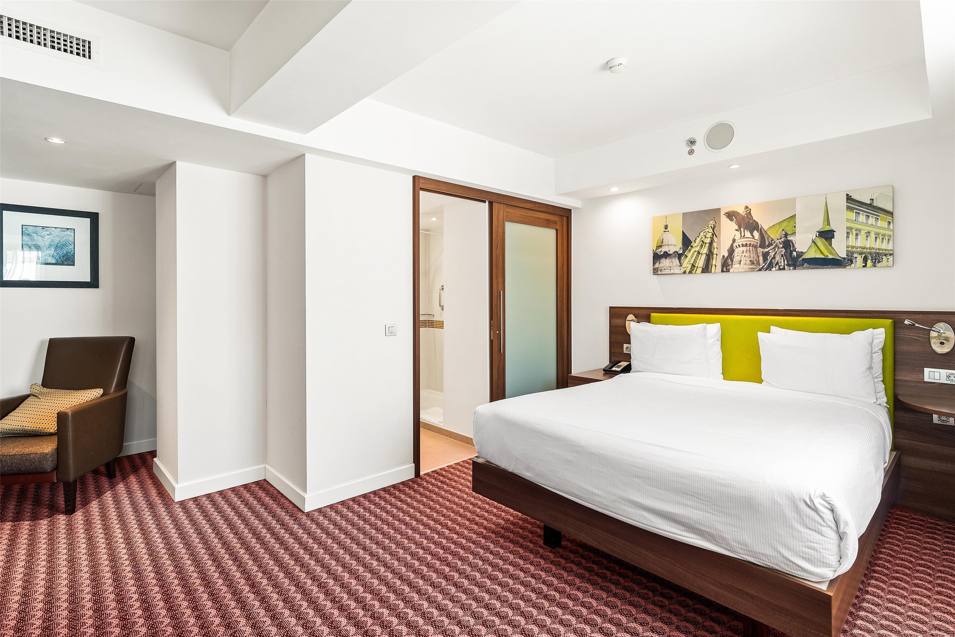 A hotel room with a bed, a chair, a painting, and a door leading to a bathroom.