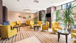 A lobby with yellow armchairs, a patterned carpet, a chandelier, a flat-screen TV, and potted plants. - Hampton by Hilton Cluj-Napoca (Cluj-Napoca)