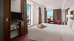 A hotel room with a large bed, a television on a wooden stand, a small table, a chair, and a view of a cityscape through large windows. - Hampton by Hilton Cluj-Napoca (Cluj-Napoca)