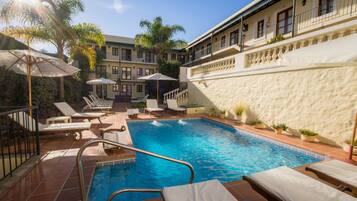 A swimming pool with sun loungers and umbrellas, surrounded by a building with balconies and potted plants.