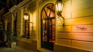 A building with a dark entrance door, a sign reading "Con Antigua," and a potted plant on the sidewalk. - Don Antonio Posada (Colonia del Sacramento)