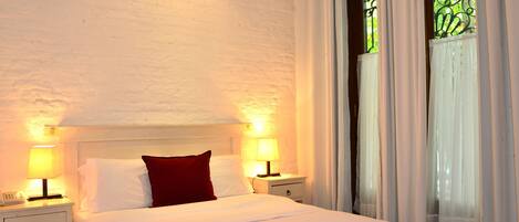 A neatly made bed with white bedding and a red pillow, two bedside lamps, a nightstand with a phone, and a window with white curtains and a decorative iron grille.