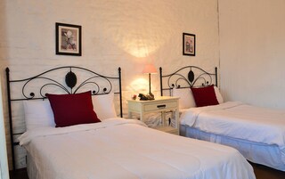 Una habitación con dos camas individuales, cada una con ropa de cama blanca y almohadas rojas. Entre las camas, un mesita de noche con una lámpara.