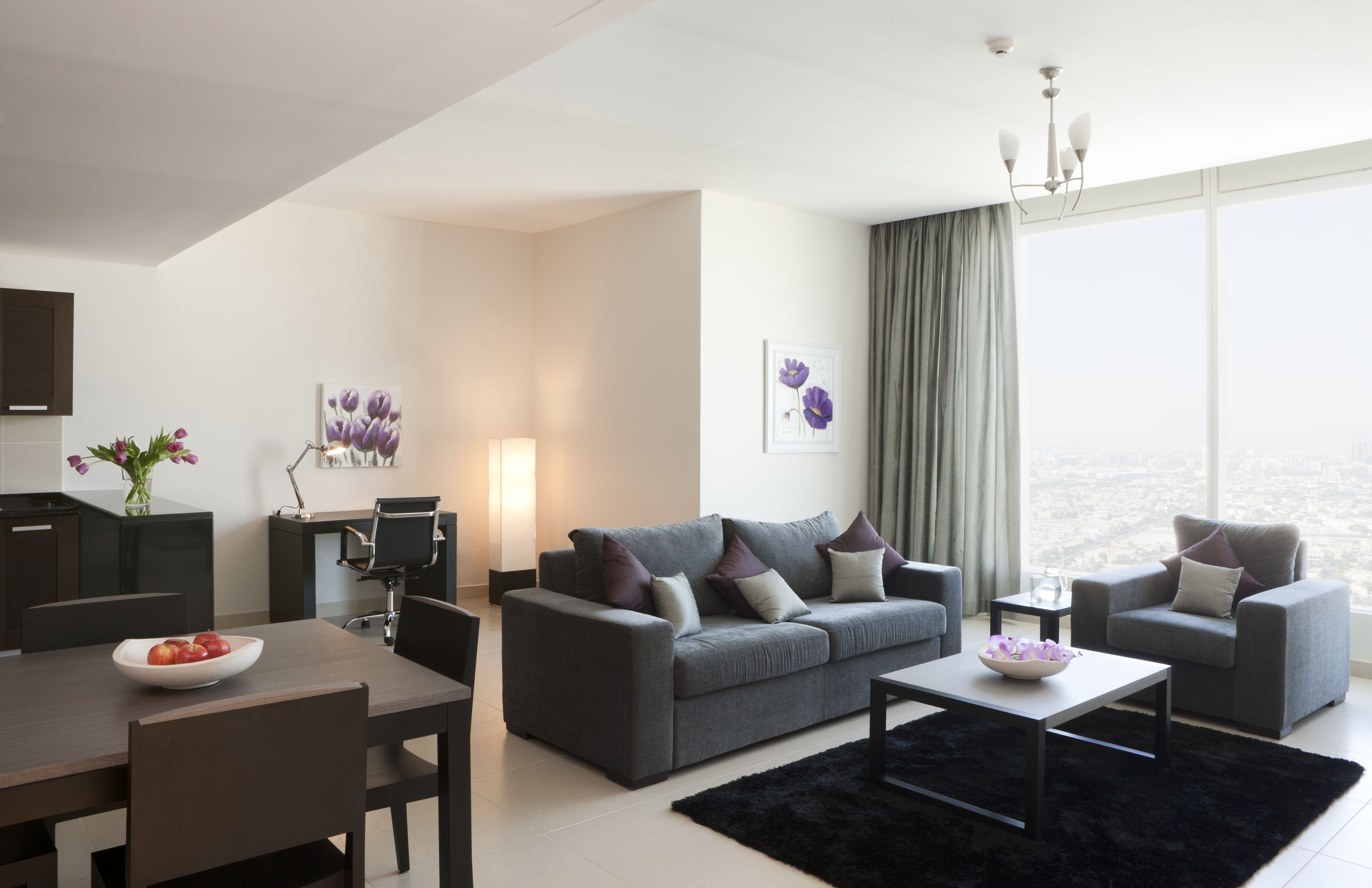 A modern living room with a grey sofa, two armchairs, a coffee table, and a dining area with a bowl of fruit.