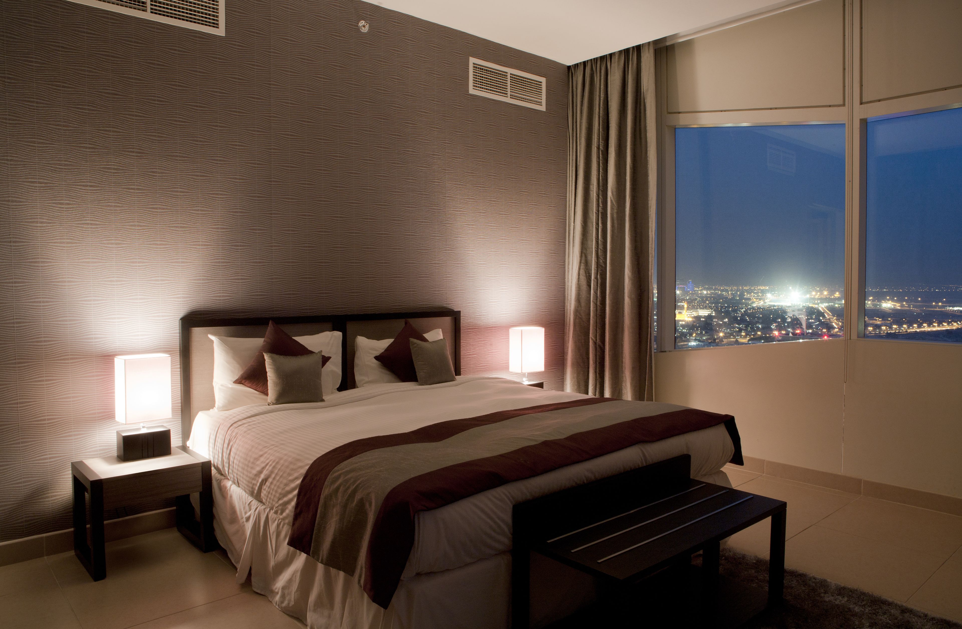 Habitación de hotel con una cama grande, dos mesitas de noche con lámparas, un banco, vista a la ciudad a través de amplias ventanas y una pared con textura.