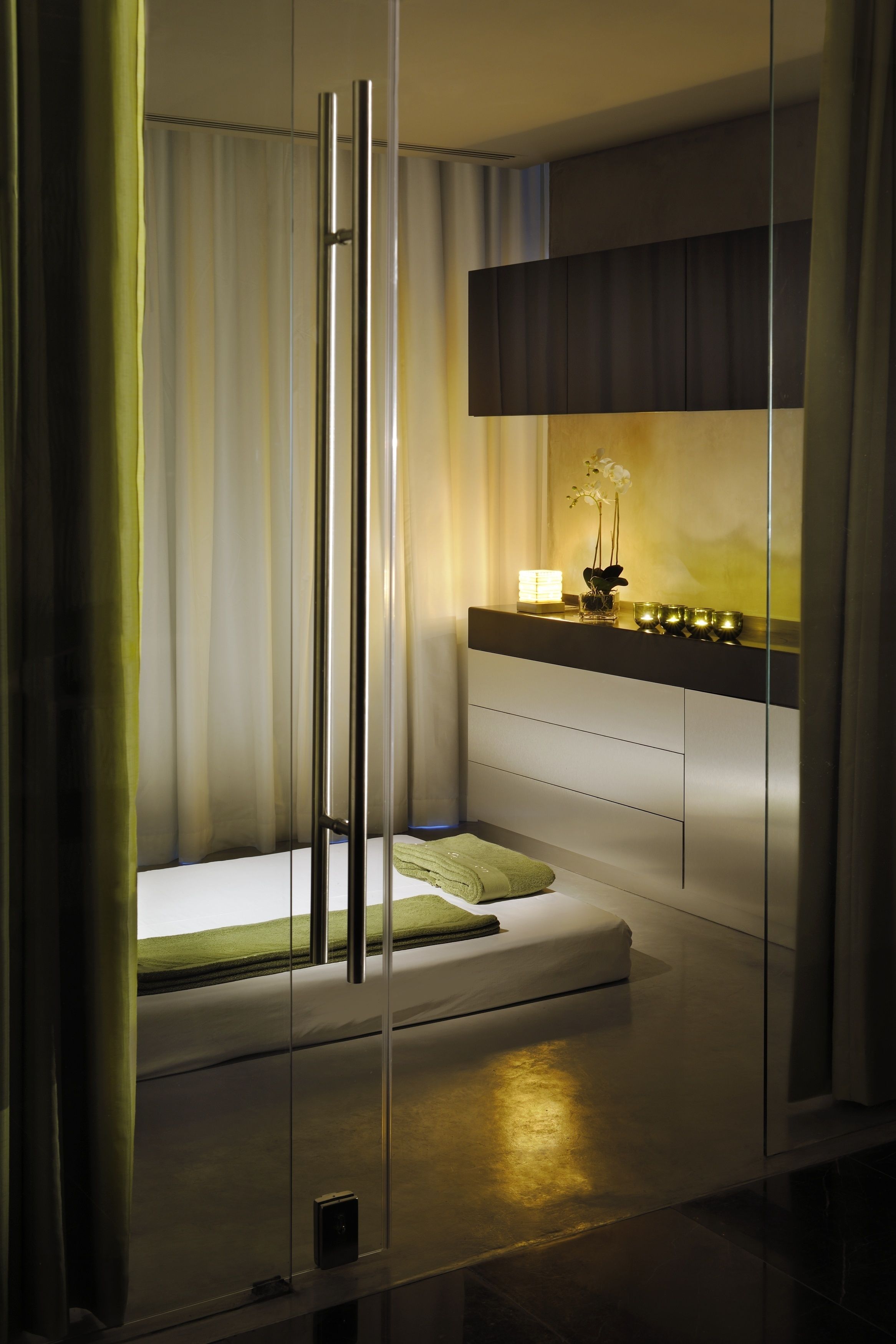 A modern bedroom with a glass partition, a bed with a green pillow, a bedside table with a lamp, and a shelf with decorative items.