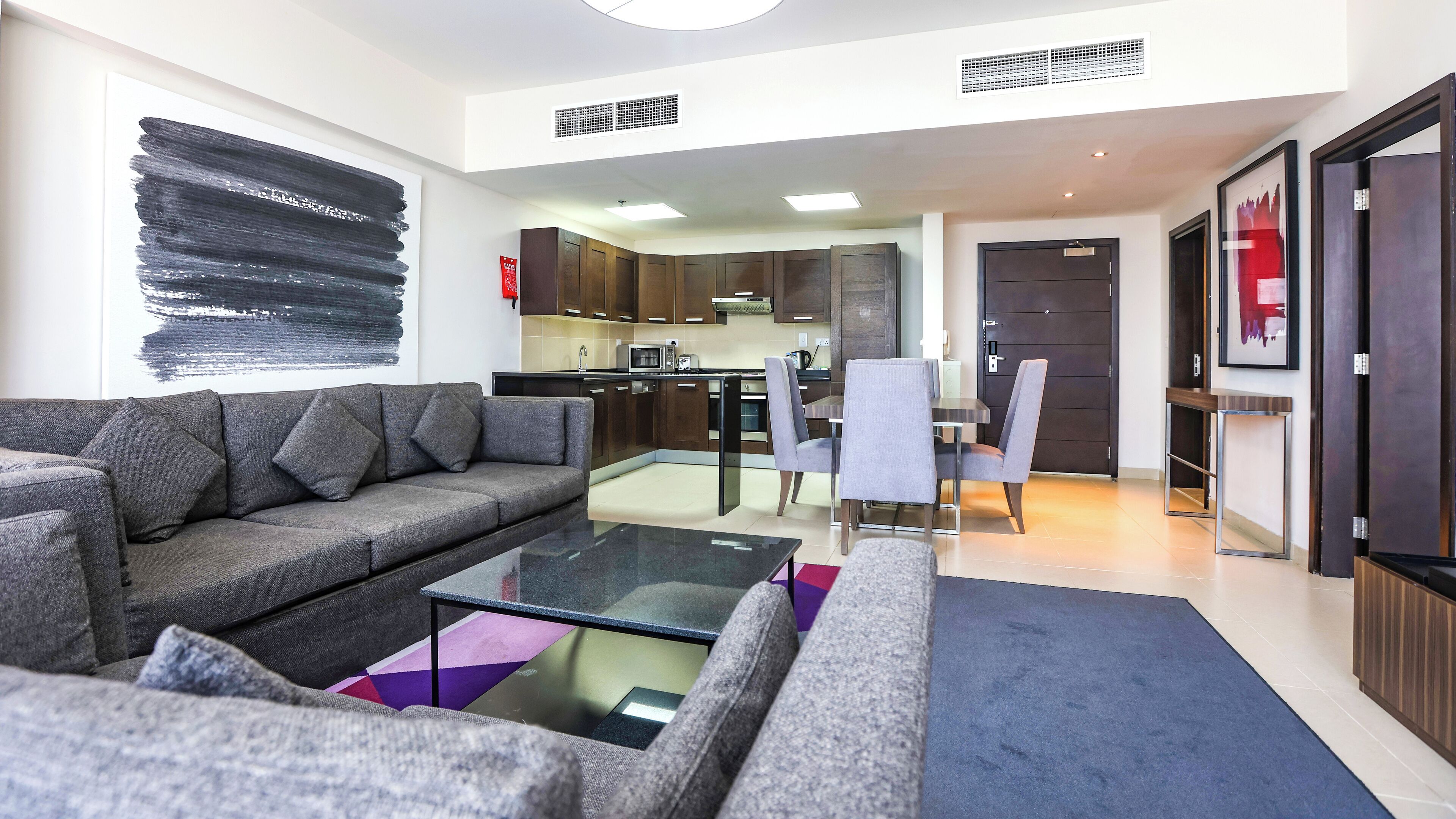 A modern living room with a grey sofa, a glass coffee table, and a dining area with grey chairs.