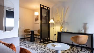 Habitació d'hotel moderna amb un sofà, una taula rodona, una cadira, un escriptori i una planta decorativa.
