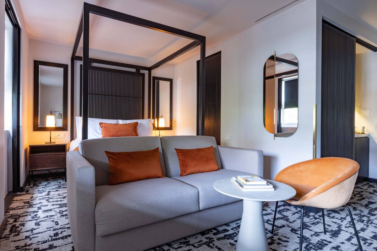 Een moderne hotelkamer met een grijze bank, oranje kussens, een ronde witte tafel en één oranje fauteuil.