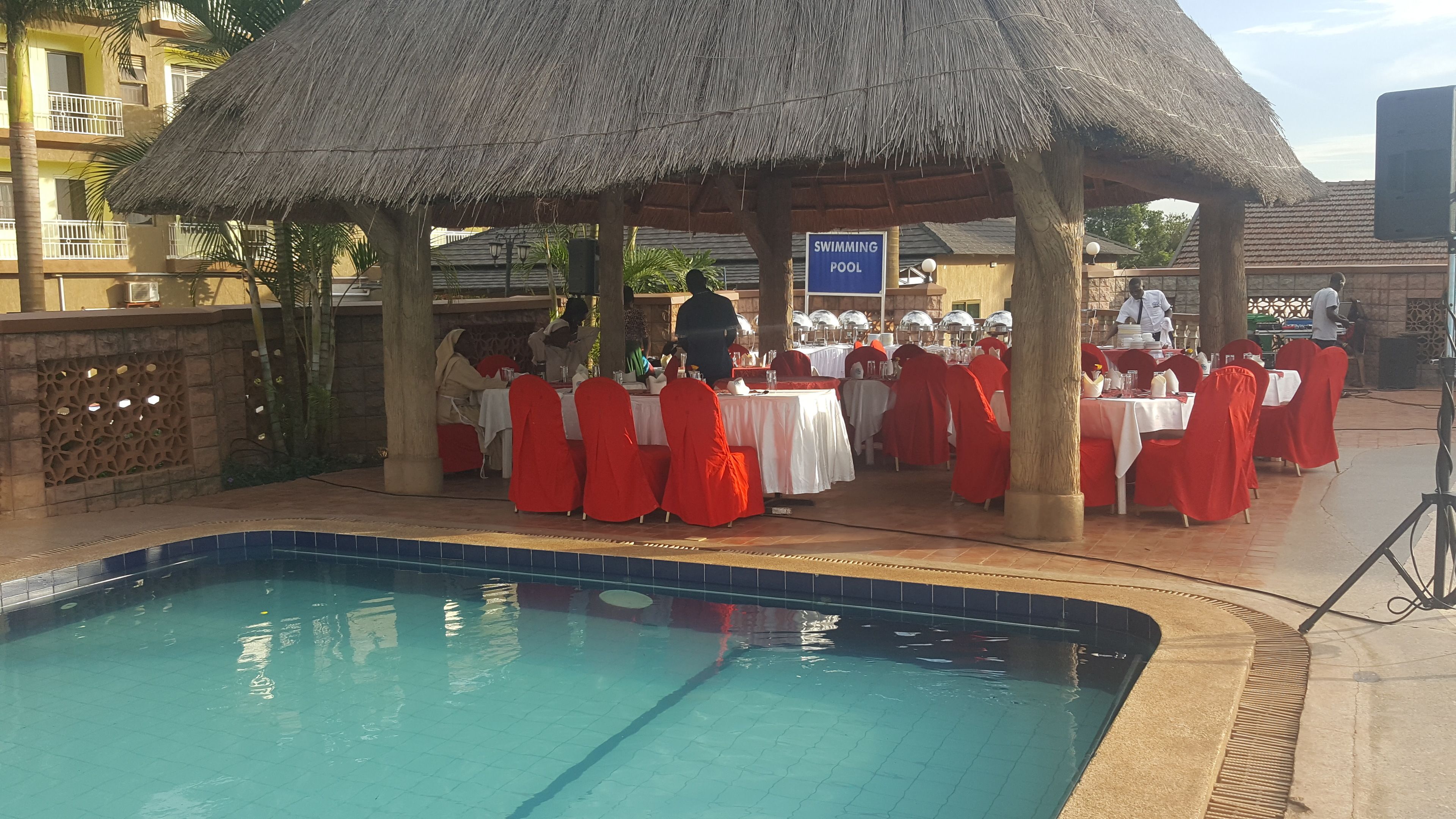Un espace de restauration aménagé au bord de la piscine, avec des tables dressées pour les clients, un panneau indiquant la présence d’une piscine et un pavillon à toit de chaume.