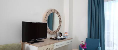 A hotel room with a television, a desk with a coffee maker, a chair, a small table with a vase, and a large window with curtains.