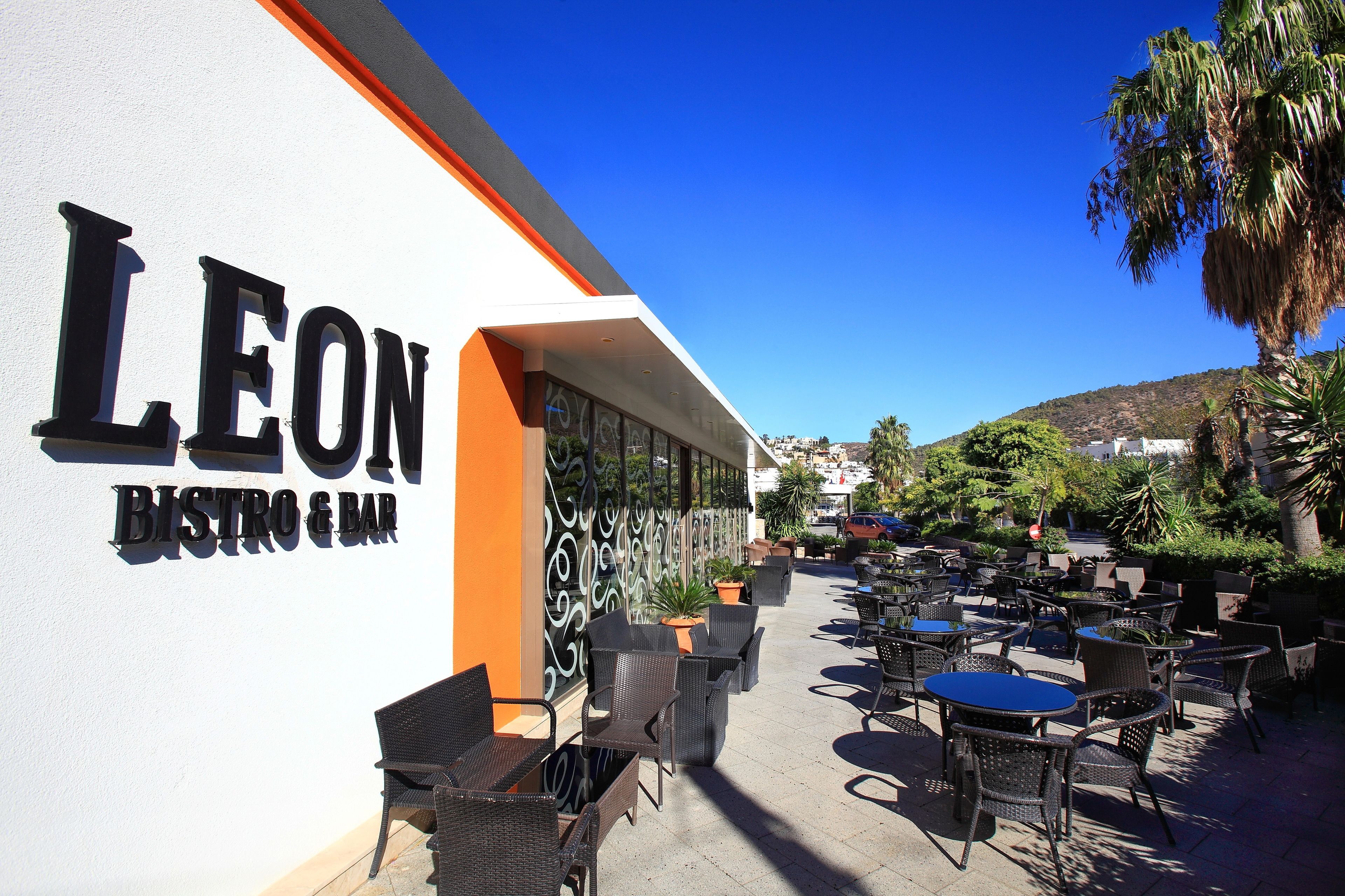 An outdoor seating area with wicker chairs and tables, a white wall with the name "LEON BISTRO & BAR" in black letters, and a clear blue sky.