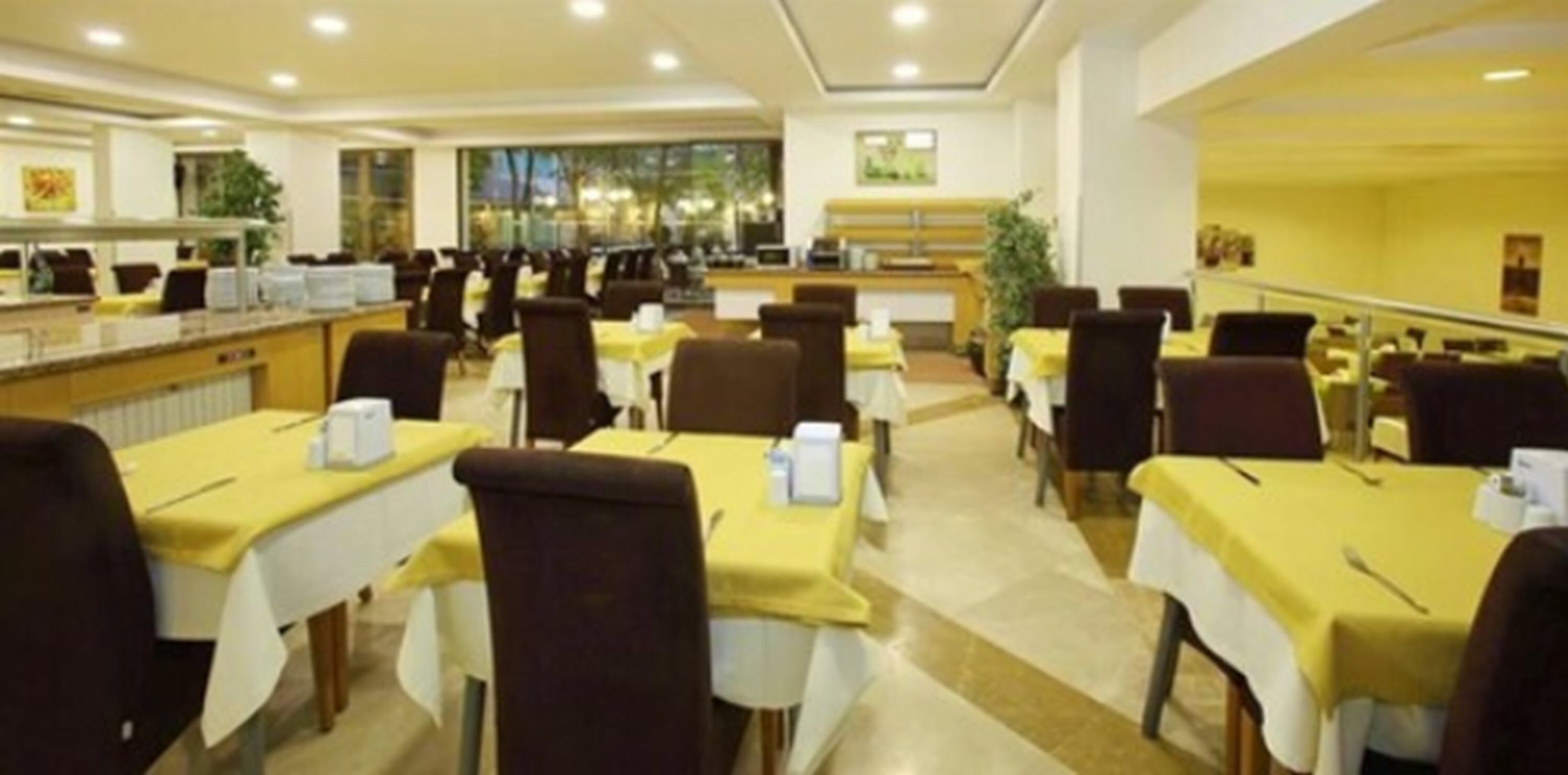 A hotel dining area with multiple tables and chairs, a counter with a cash register, and a view of the outdoors through large windows.