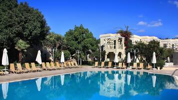 A swimming pool with lounge chairs and umbrellas, surrounded by trees and a building with a white facade.