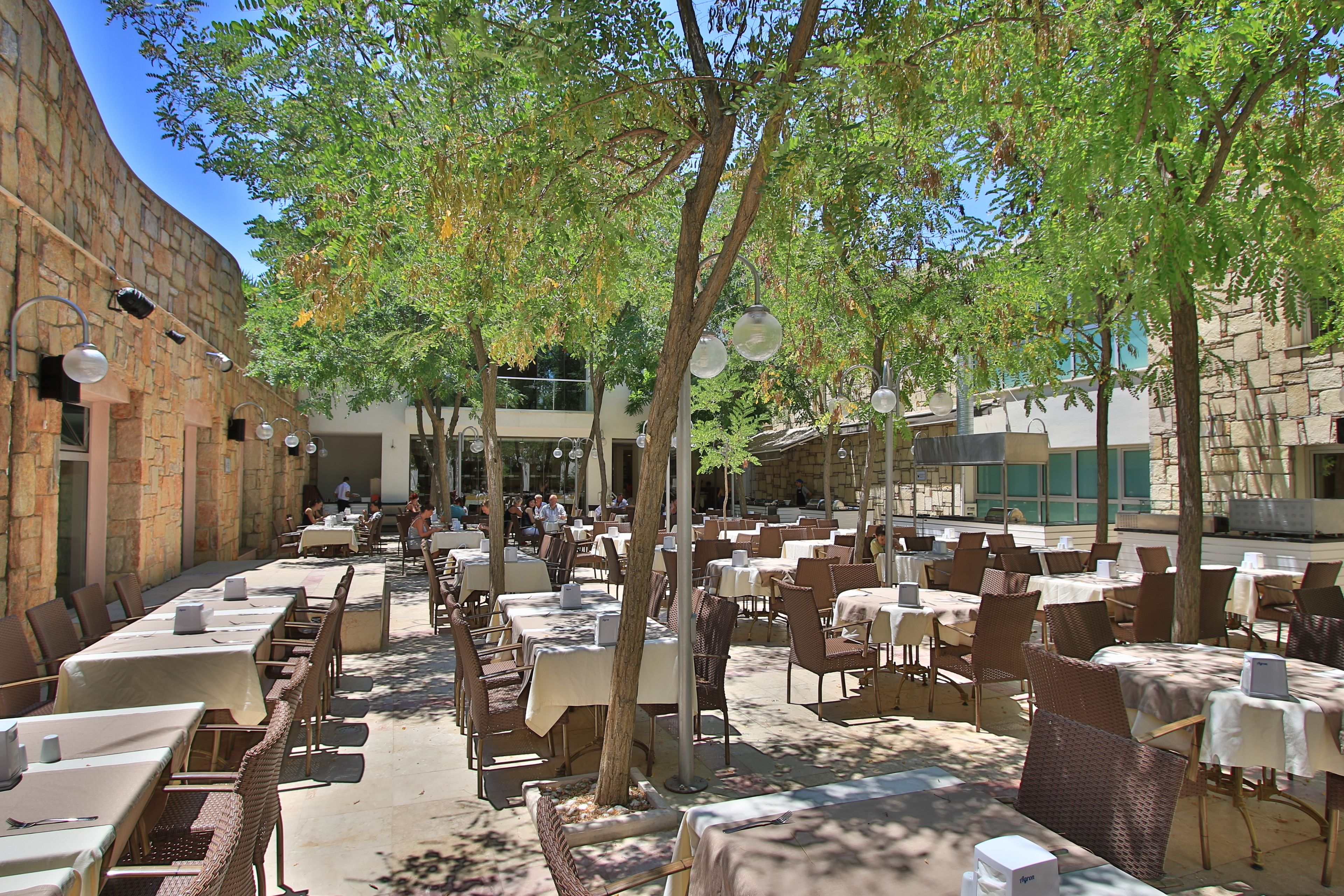 An outdoor dining area with multiple tables and chairs, surrounded by trees and stone walls.
