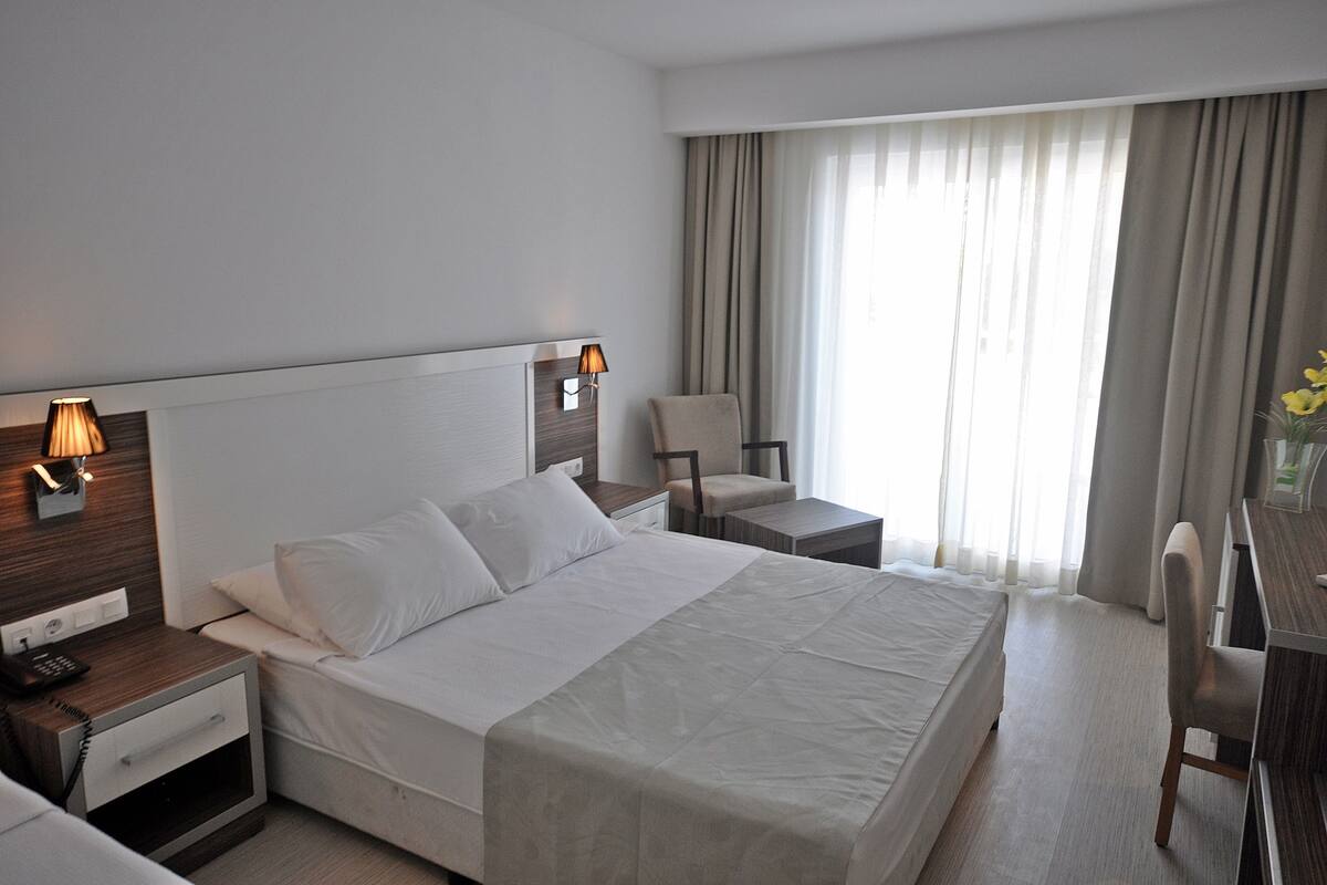 A hotel room with a large bed, a desk, a chair, a nightstand with a lamp, and a window with curtains.