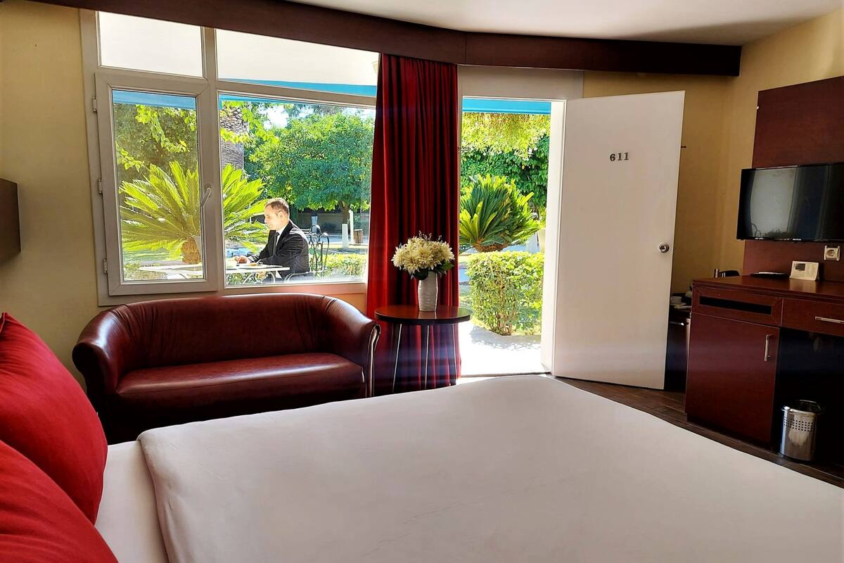 Una camera d'albergo con un letto grande, una poltrona in pelle, una scrivania con una TV e vista su un giardino tramite una porta in vetro.
