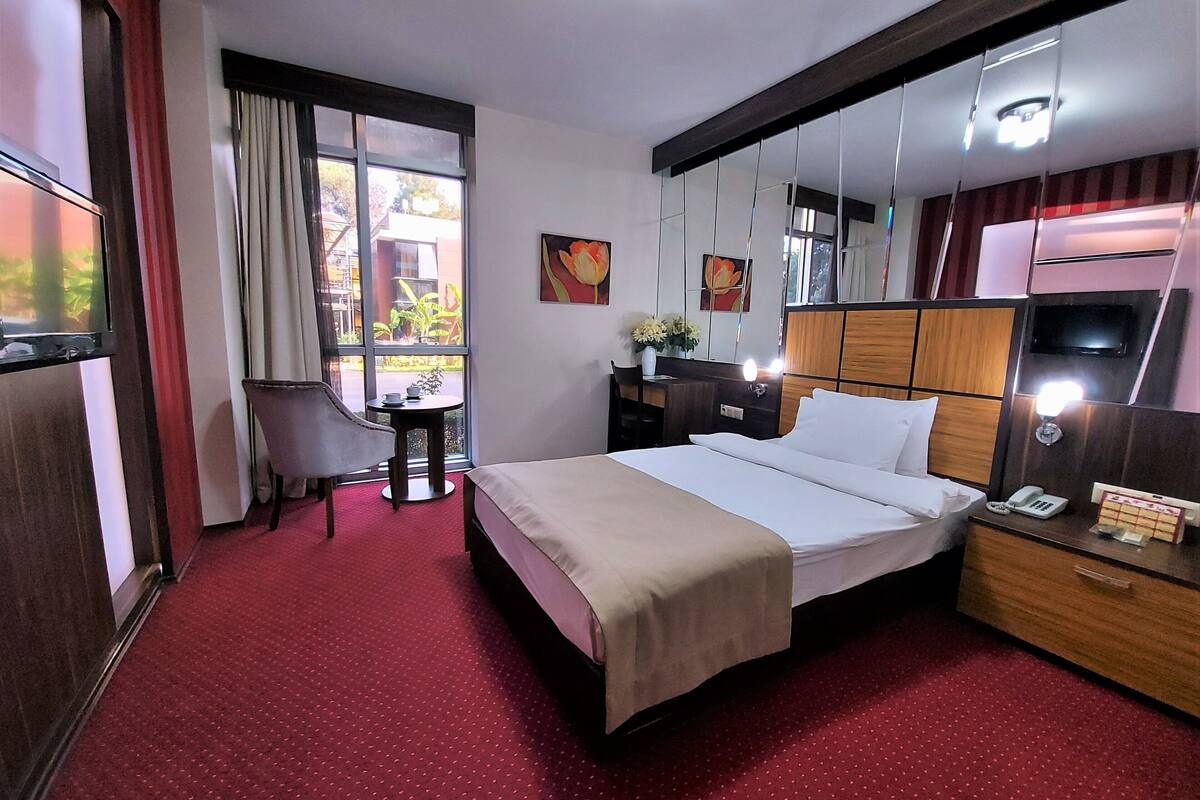 Una camera d'albergo con un letto, una scrivania, una sedia, una televisione e un'ampia finestra con tende.