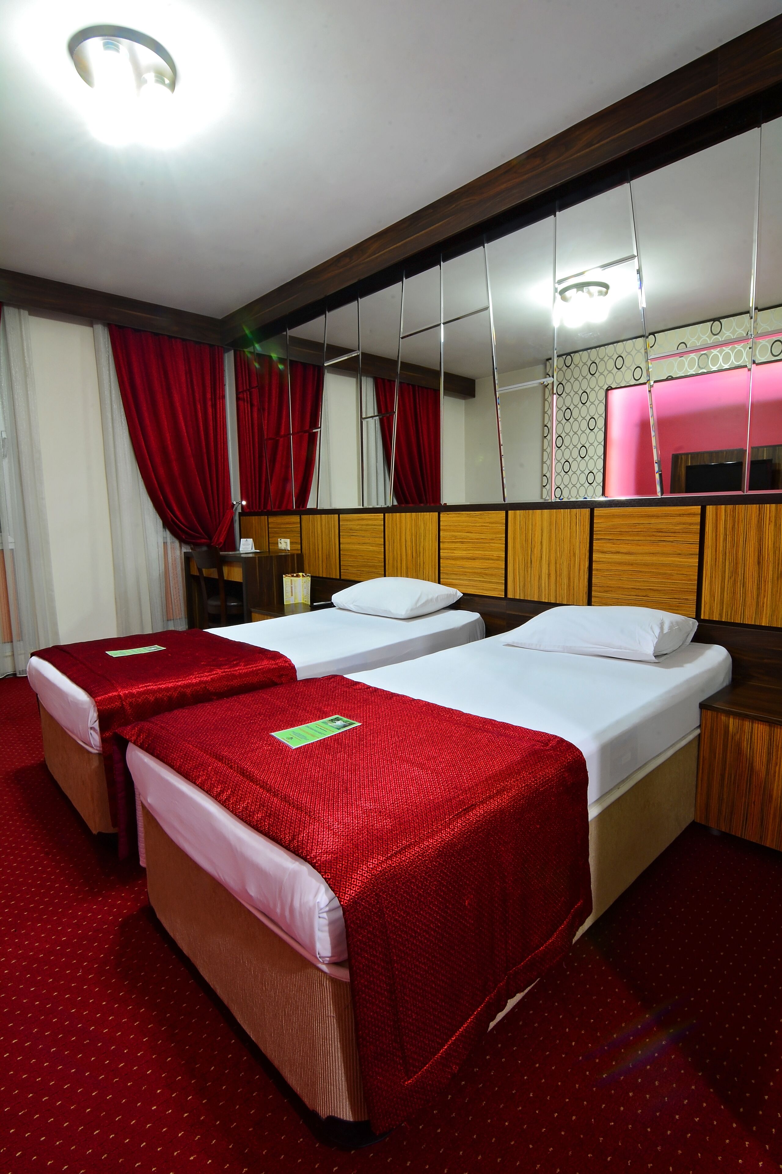 Une chambre d’hôtel avec deux lits, des rideaux rouges, une tête de lit en bois, un miroir et une petite table avec une lampe.