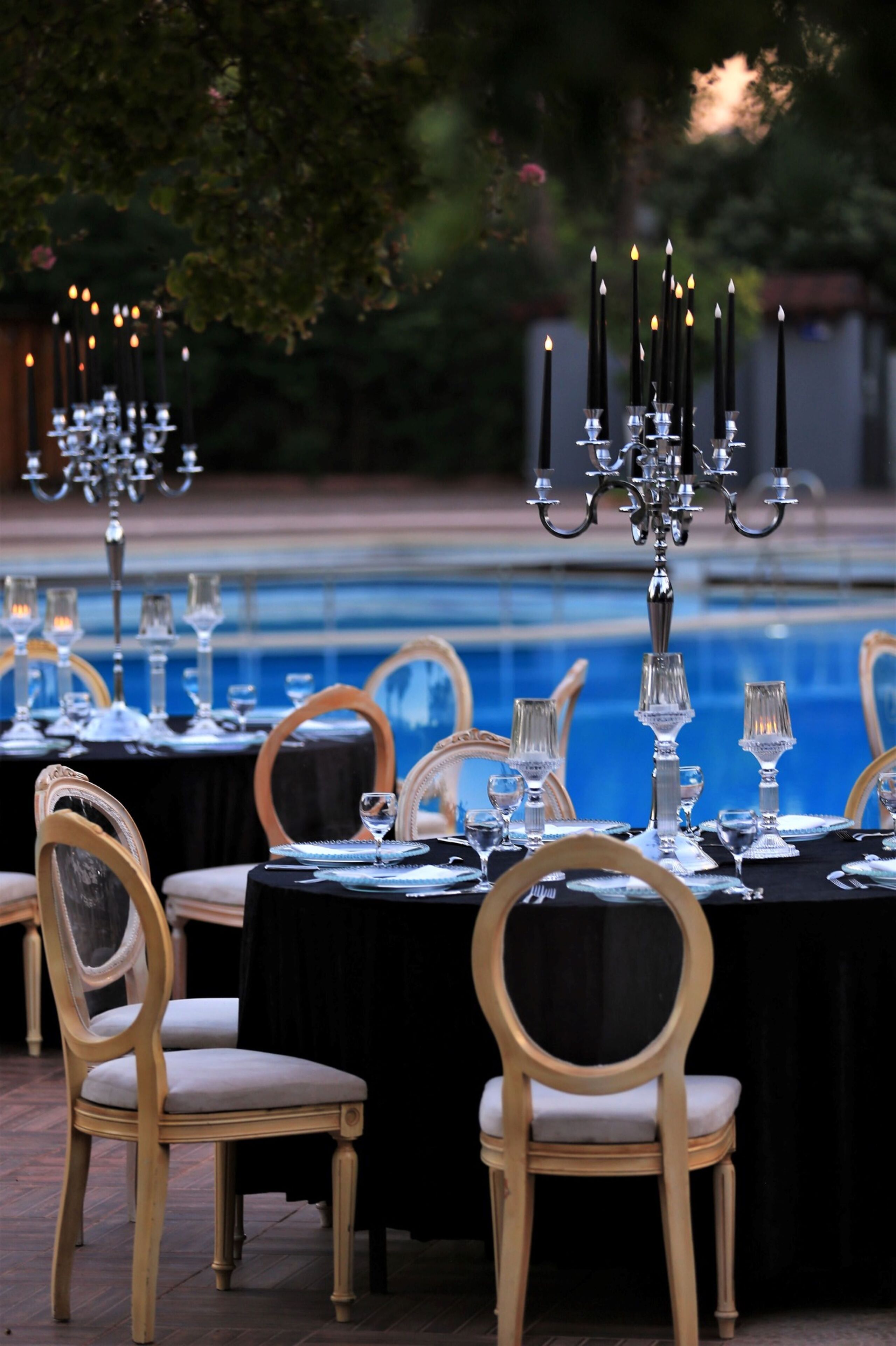 Un área de comedor al aire libre con mesas dispuestas para una comida, rodeada de sillas y una piscina al fondo.