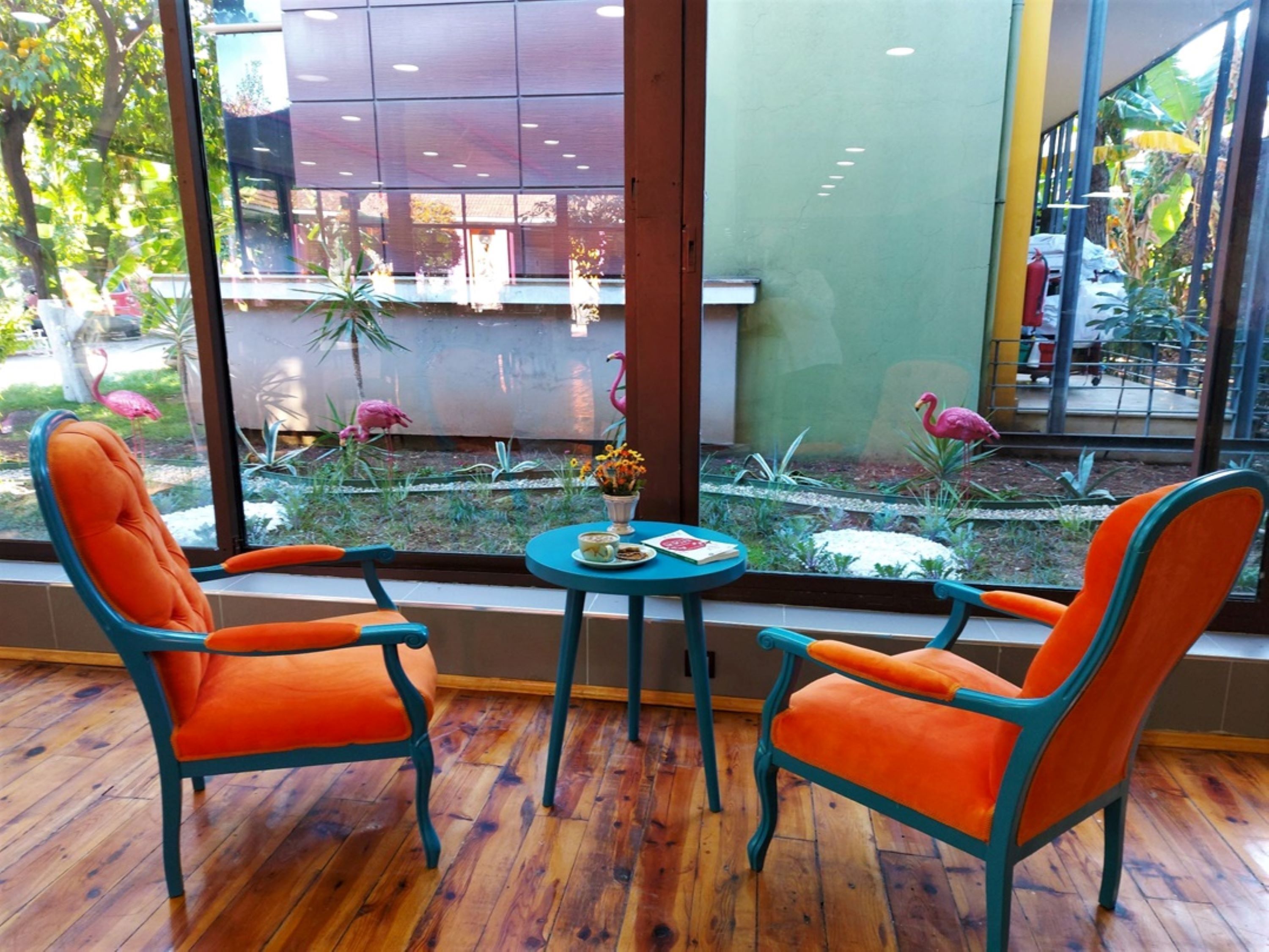 Ein Zimmer mit zwei orangen Sesseln und einem kleinen Tisch, große Fenster mit Blick auf ein Gebäude und Pflanzen draußen.