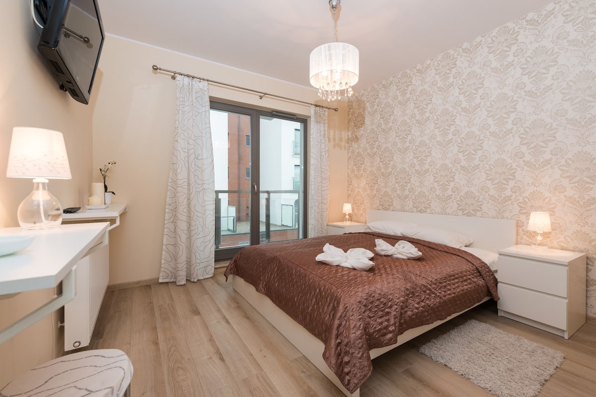 Quarto com cama, mesas de cabeceira, televisão, janela com cortinas e um candelabro.
