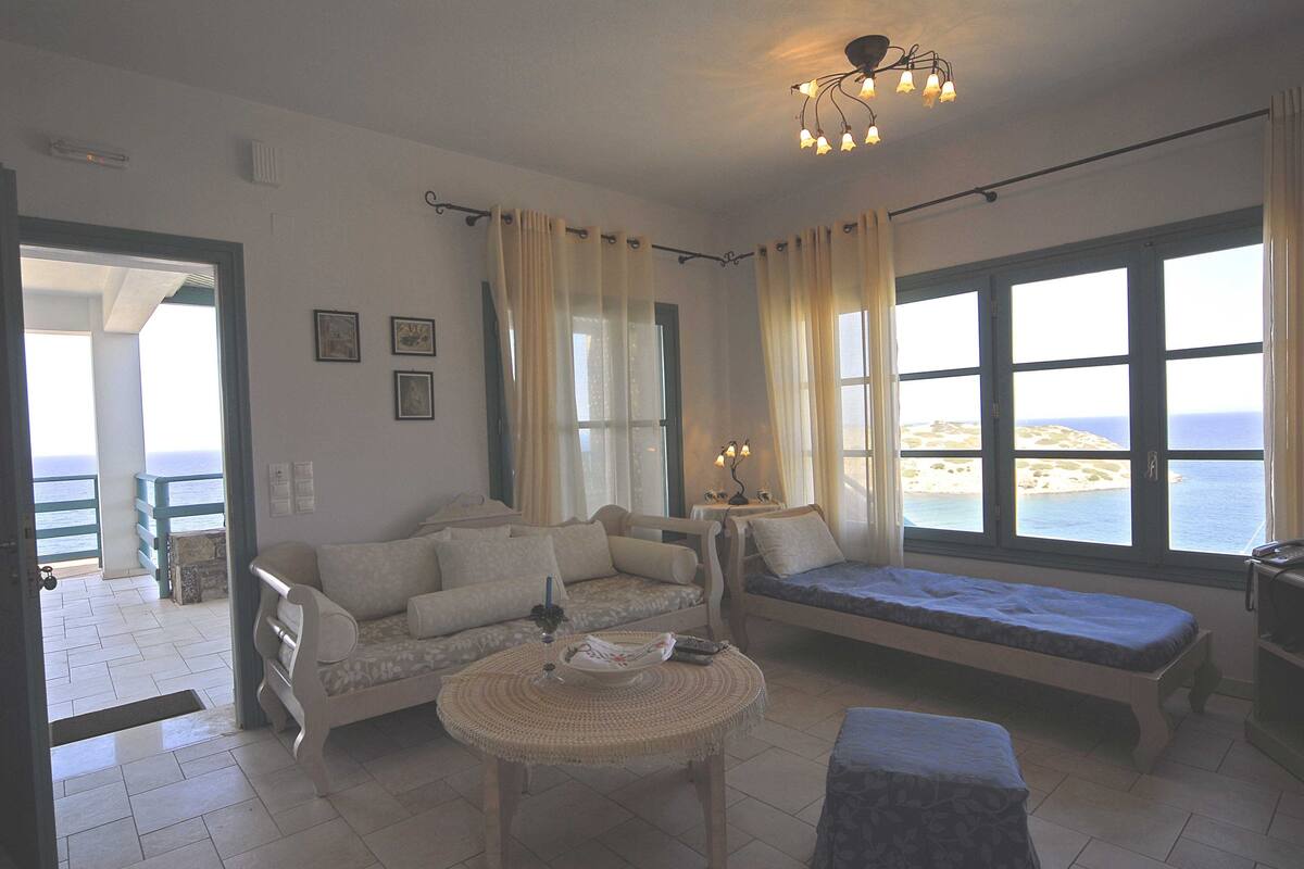 A room with a sofa, a bench, a small table, and large windows with a view of the sea.