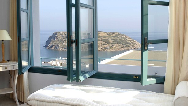 A room with a bed, a table with glasses, and a view of the sea through a window. - Mohlos Villas (Sitia)