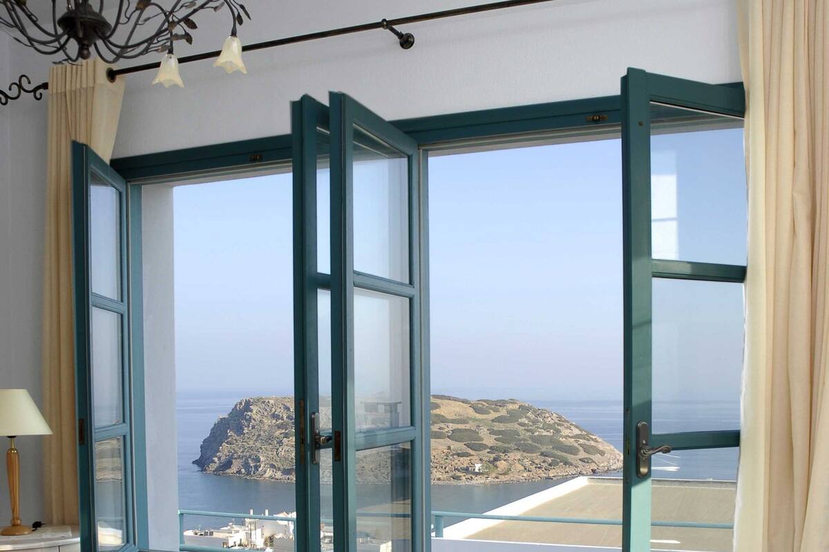A room with a bed, a table with glasses, and a view of the sea through a window.
