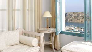 A room with a bed, a sofa, a table with a lamp, and a view of the sea through an open window. - Mohlos Villas (Sitia)