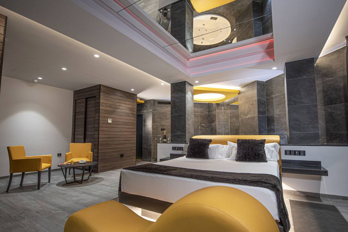 Una habitación de hotel moderna con una cama grande, una zona de estar con sillas amarillas, una mesita y un balcón acristalado con vistas.