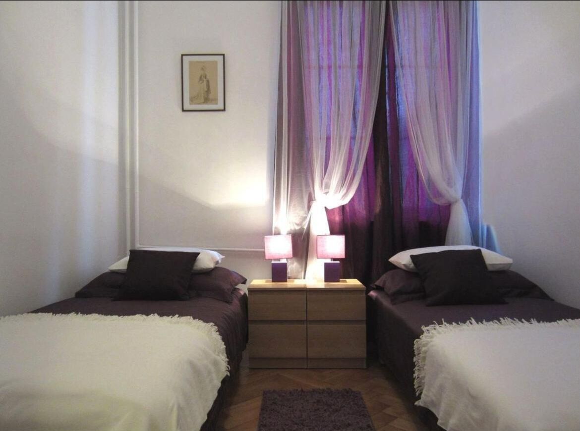 Ein Zimmer mit zwei Einzelbetten, einem Nachttisch mit zwei Lampen und einem Fenster mit durchsichtigen Vorhängen.